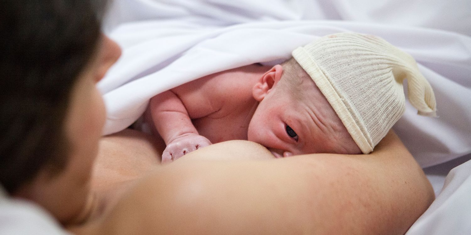 Ein Neugeborenes mit Mütze trinkt an der Brust seiner Mutter, die im Bett liegt.. Artikel "Kolostrum: Superfood für die ersten Lebenstage" öffnen.