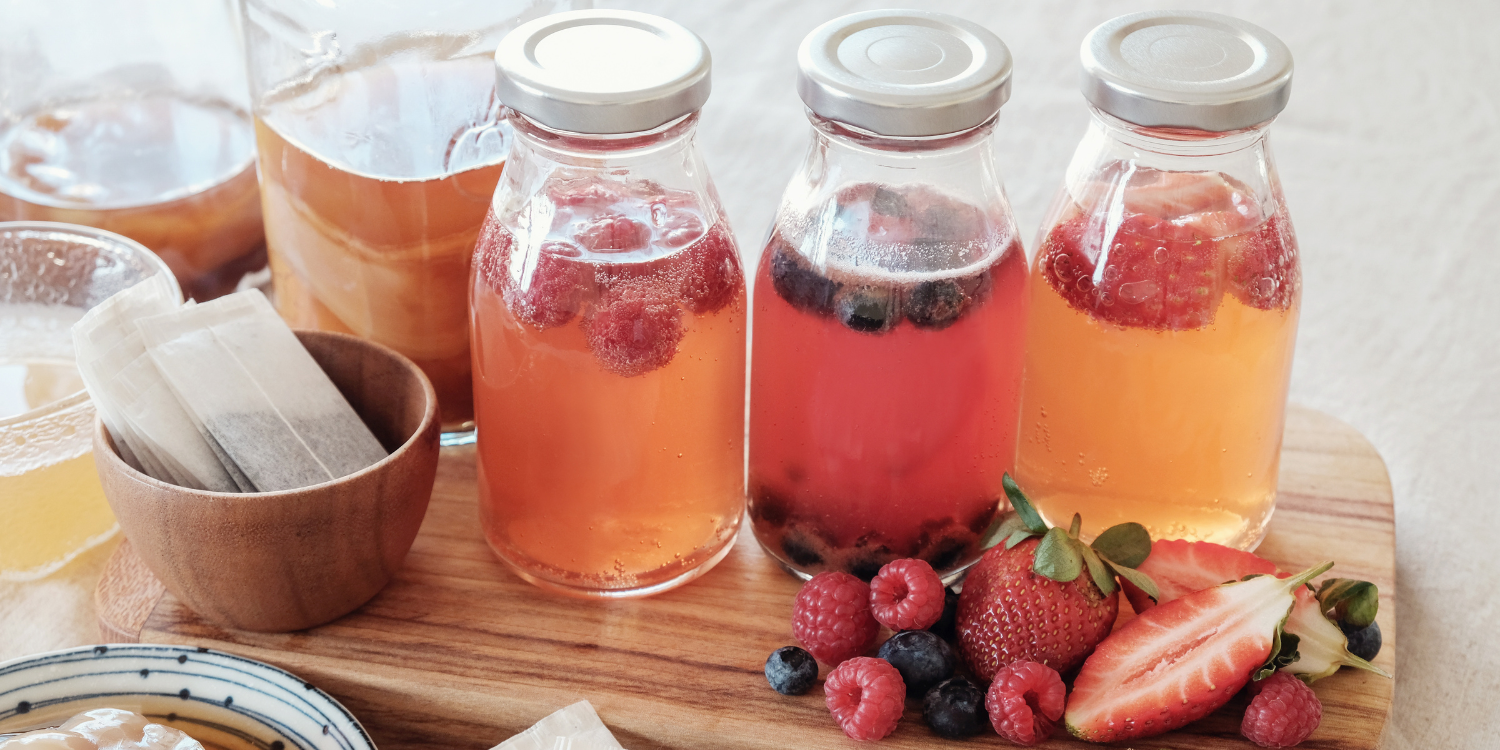 Auf einem Holzbrettchen stehen drei Schraubdeckelfläschchen mit einer sprudelnden klaren Flüssigkeit und Himbeeren, Blaubeeren und Erdbeeren. Daneben liegen einzelne Früchte und Teebeutel. . Artikel "Sommerdrinks ohne Alkohol" öffnen.