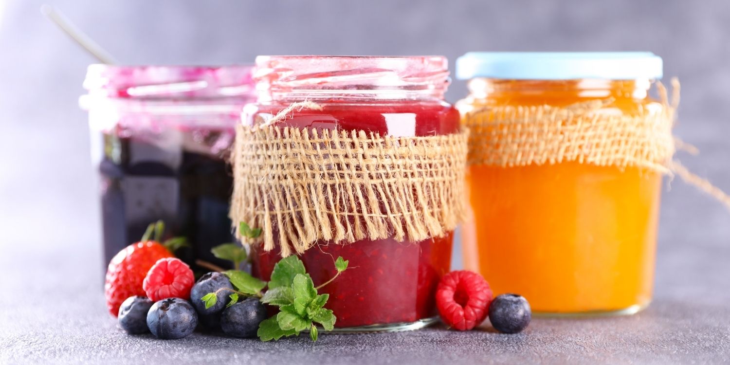 Vor einem grauen Hintergrund stehen drei Gläser mit Marmelade in Gelb, Rot und Lila, daneben liegen einige Beeren und Minzblätter.. Artikel "Marmelade darf jetzt auch offiziell Marmelade heißen" öffnen.