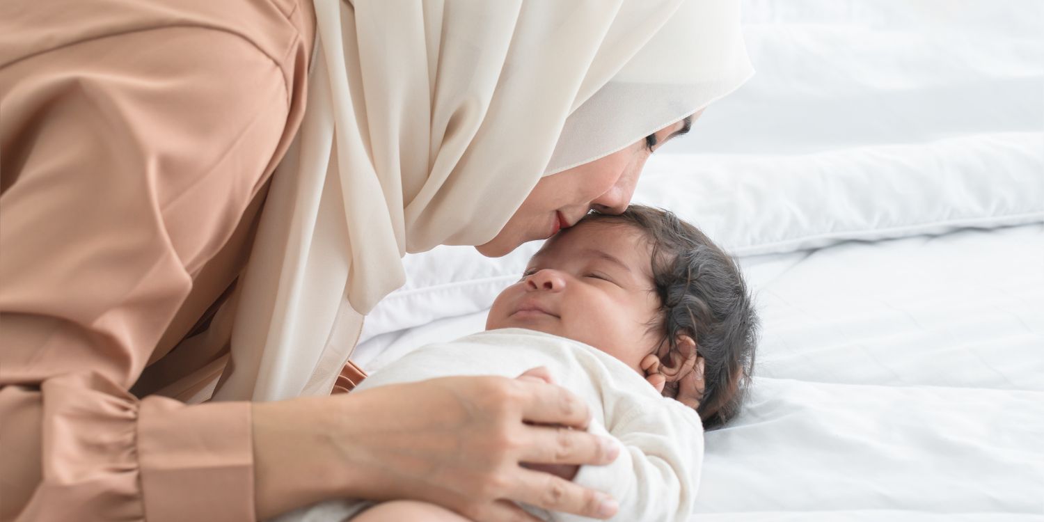 Eine junge Muslimin mit Hijab küsst ihr lächelndes, einmonatiges Neugeborenes auf die Stirn, während das Baby im Bett schläft.. Artikel "Junge Familien mehrsprachig beraten " öffnen.