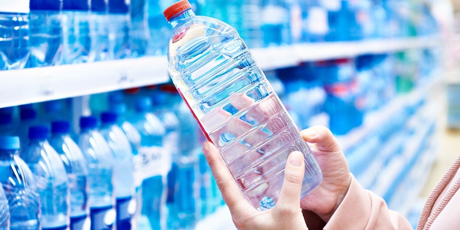 Vor einem Getränkeregal mit Wasserflaschen hält eine Frau eine PET-Flasche mit Mineralwasser in den Händen.. Artikel "Wie lange ist Mineralwasser haltbar?" öffnen.