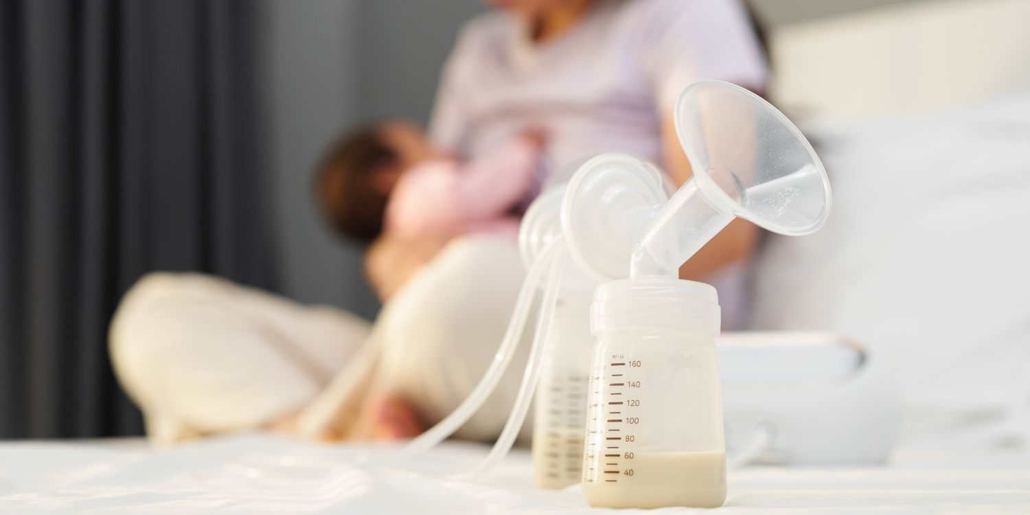 Auf einem Tisch steht eine Muttermilchpumpe mit Flasche und Inhalt. Im Hintergrund stillt eine Mutter ihr Baby.. Artikel "Muttermilch abpumpen" öffnen.