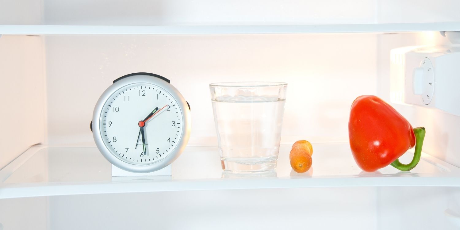 In einem Kühlschrank stehen ein Wecker und ein Glas Wasser, daneben liegen eine Möhre und eine rote Paprika.. Artikel "Fette besser zum Frühstück oder zum Abendbrot essen?" öffnen.