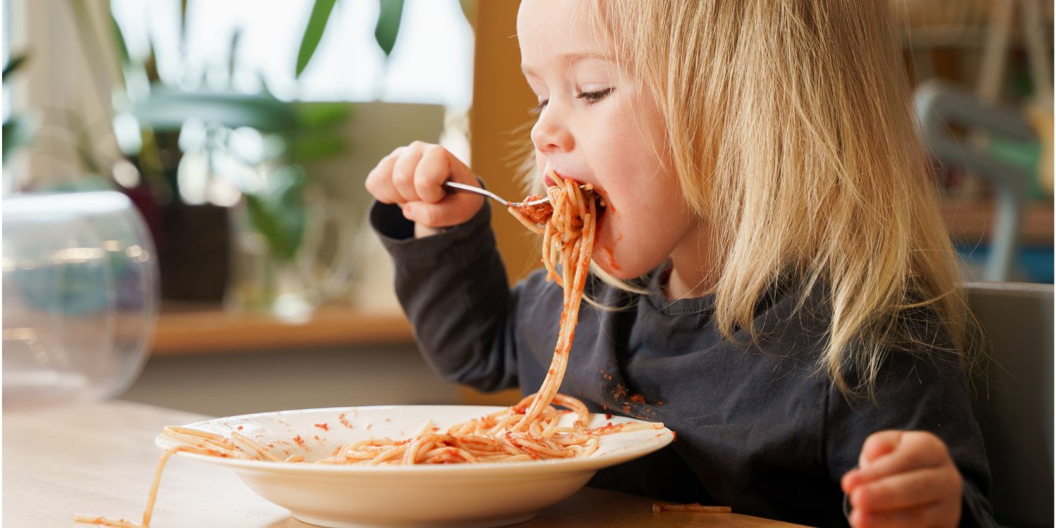 Ein blondes Mädchen im Kleinkindalter sitzt an einem Esstisch und schiebt sich eine volle Gabel mit Spaghetti in den Mund.. Artikel "Ist mein Kind gut versorgt?" öffnen.