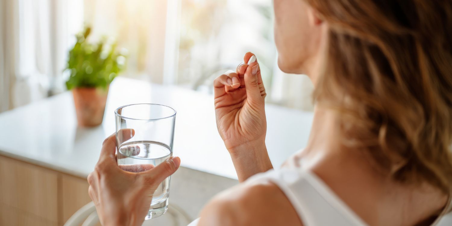 Von hinten gesehen hält eine Frau mit welligem Haar eine weiße Kapsel an ihre Lippen, während sie ein durchsichtiges Glas Wasser in der Hand hält.. Artikel "Wann Nahrungsergänzungsmittel sinnvoll sind" öffnen.