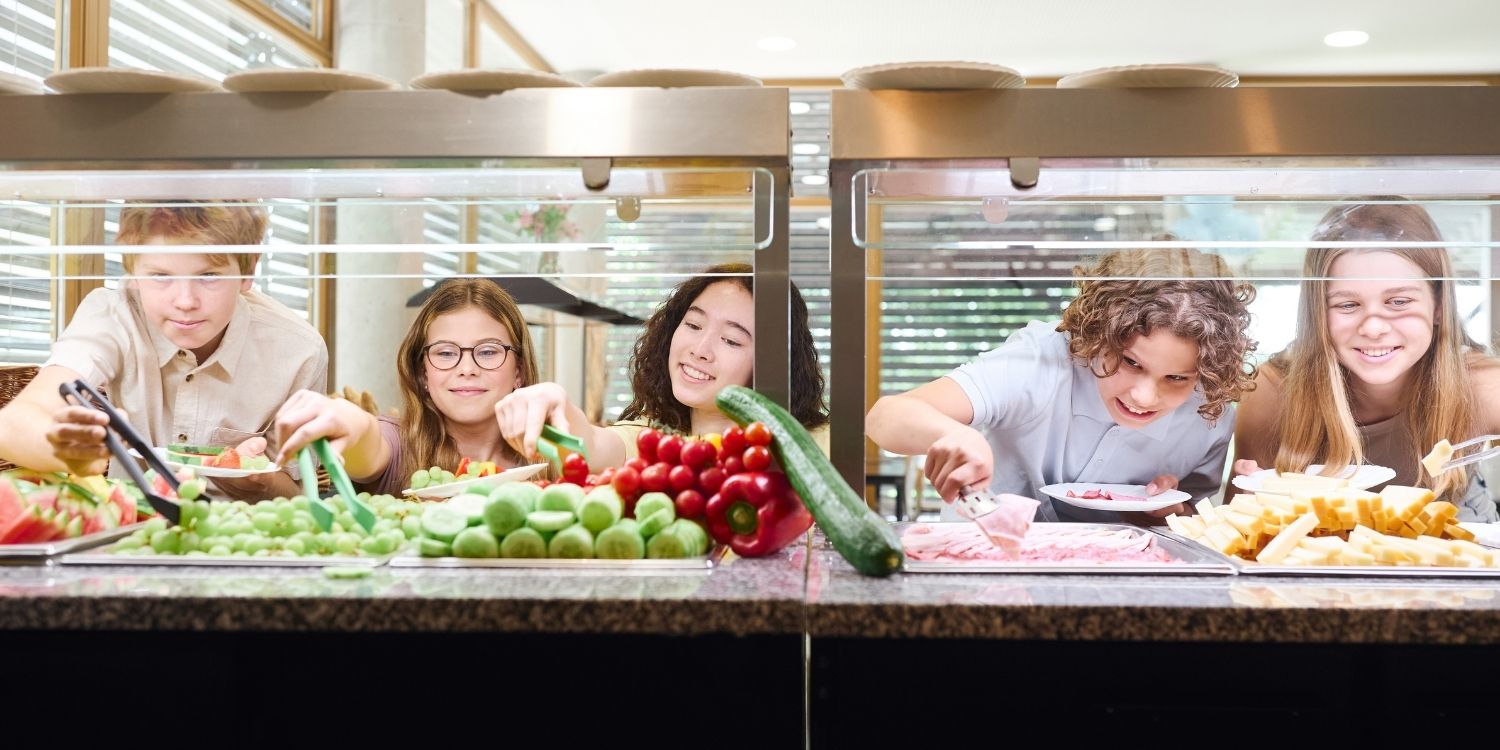 Drei Mädchen und zwei Jungen im Teenageralter bedienen sich an einer Essensausgabe mit frischem Obst und Gemüse.. Artikel "Impulse für gesundes Essen" öffnen.