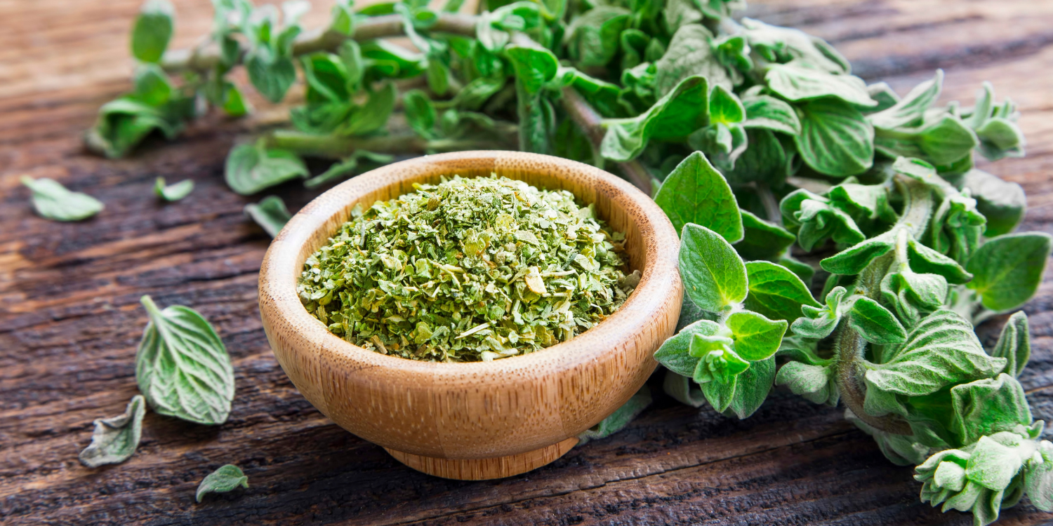 Auf einem dunklen Holztisch steht ein Schälchen mit getrocknetem, zerkleinertem Oregano. Daneben liegen frische Zweige.. Artikel "Oregano vielseitig kombinieren" öffnen.