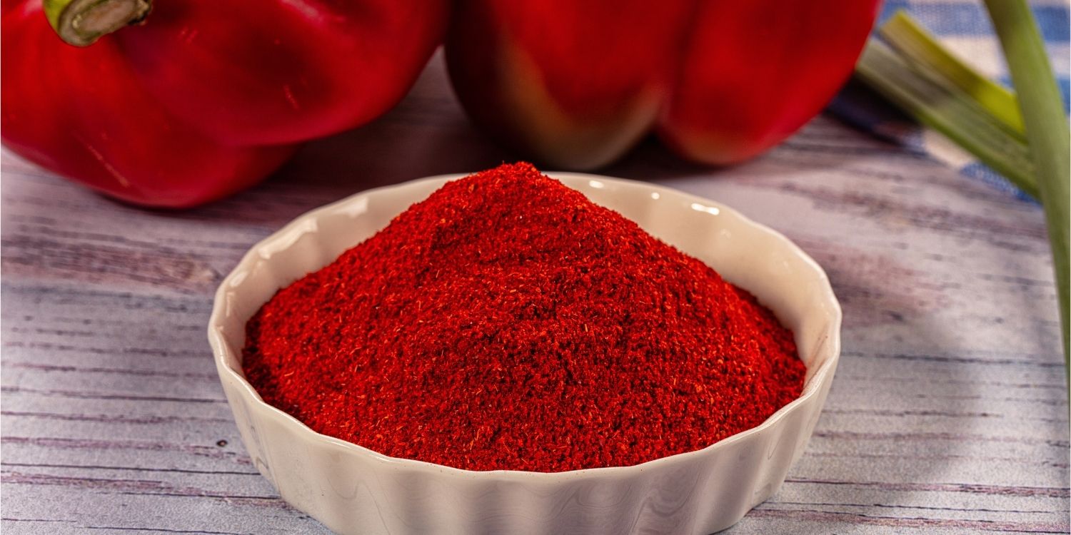 Auf einem Holztisch steht eine weiße Porzellanschüssel mit gemahlenem Paprikapulver. Im Hintergrund liegen zwei rote Paprikaschoten.. Artikel "Paprikapulver – von mild bis scharf" öffnen.