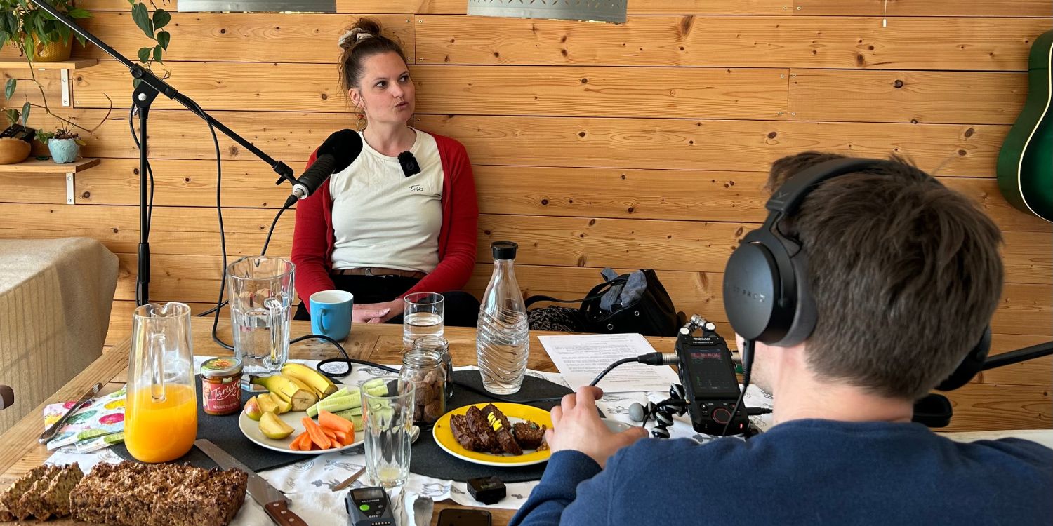 Für eine Podcast-Aufnahme sitzt ein Mann mit Kopfhörer und Mikro einer dunkelhaarigen Frau an einem Tisch gegenüber auf dem ein Imbiss steht. . Artikel "Alltagsgeschichten über Ernährung, Nachhaltigkeit und Zukunft " öffnen.