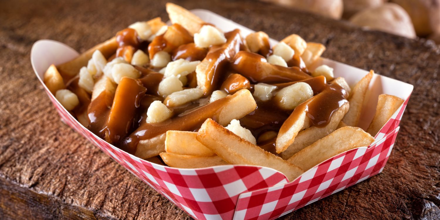 Auf einem groben Holztisch steht eine rot-weiß karierte Pappschachtel mit Pommes frites, die mit Bratensoße und Käse bedeckt sind.. Artikel "Kanada auf dem Teller" öffnen.