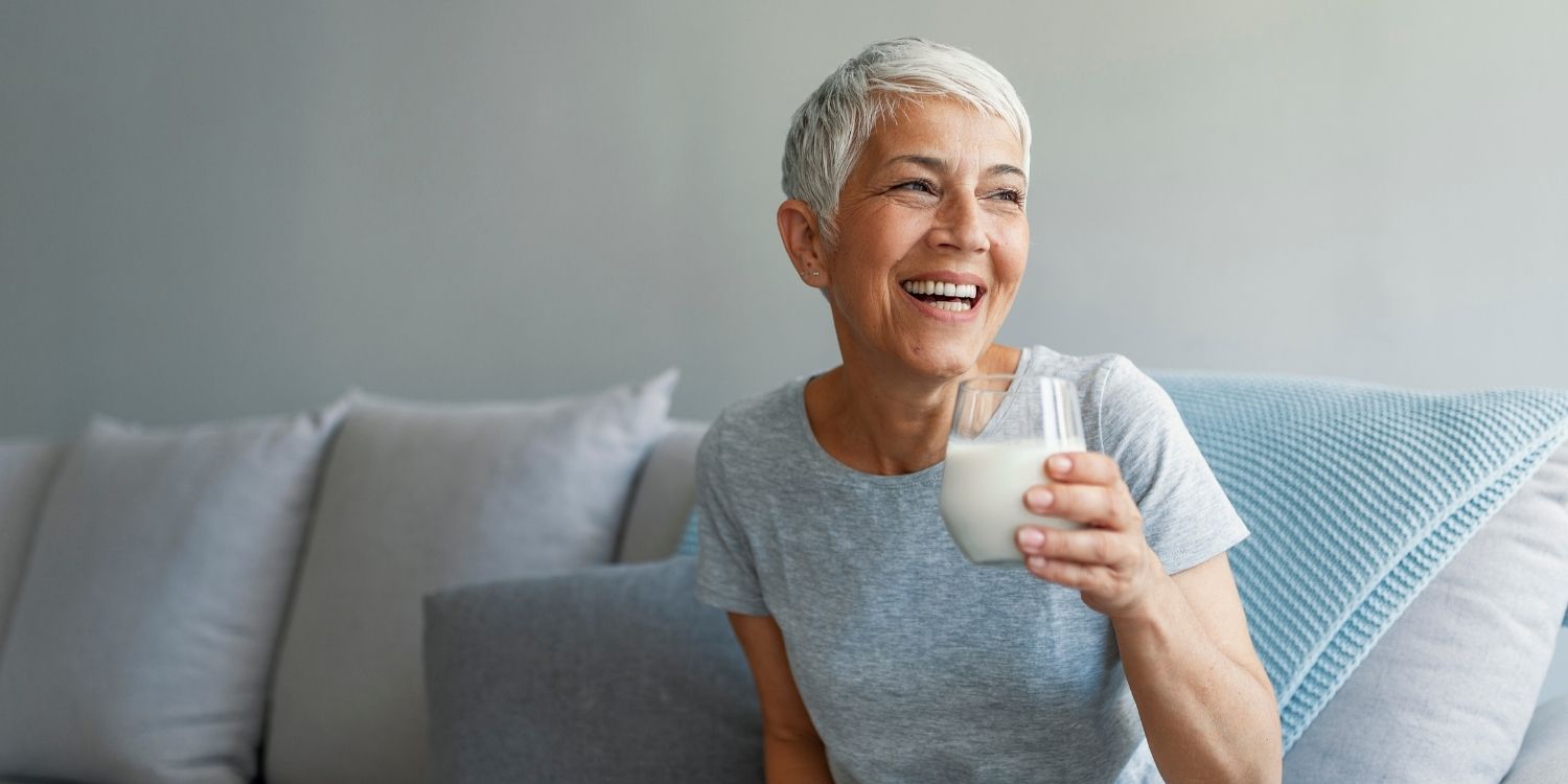 Eine Frau mit kurzen grauen Haaren sitzt lachend auf einem Sofa und hält ein Glas Milch in der Hand. . Artikel "Anders essen in den Wechseljahren" öffnen.
