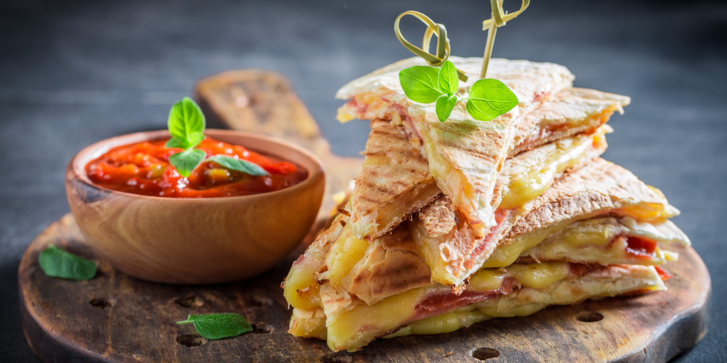 Auf einem Holzbrett liegen in Dreiecke geschnittene Quesadillas übereinander. Daneben steht ein Schüsselchen mit roter Salsa.. Artikel "Mexiko auf dem Teller" öffnen.