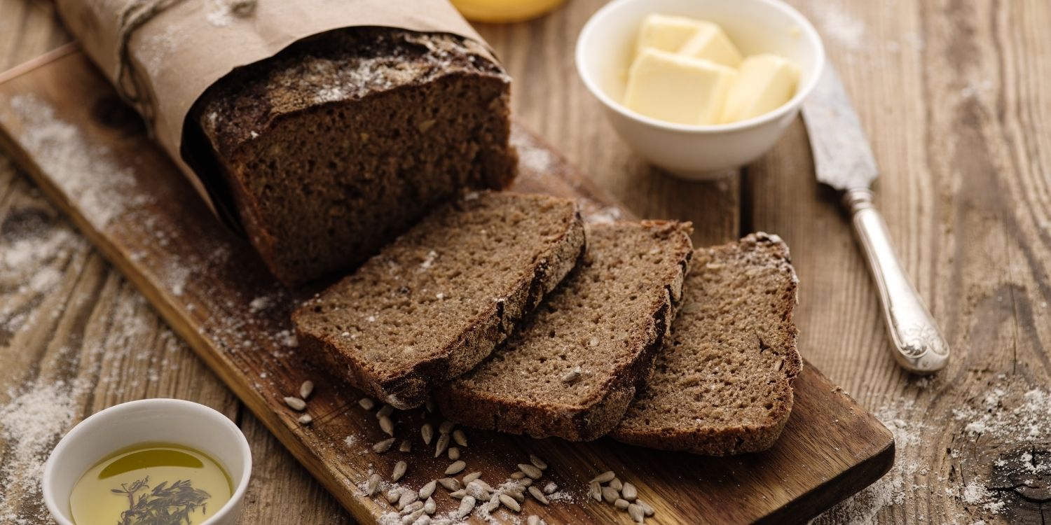 Auf einem Holzbrett liegt ein eckiges dunkles Roggenbrot mit Papiermanschette, von dem 3 Scheiben abgeschnitten sind. Daneben sind Butter, ein Messer und ein Schälchen mit Honig.. Artikel "Roggenbrot ist das Brot des Jahres " öffnen.