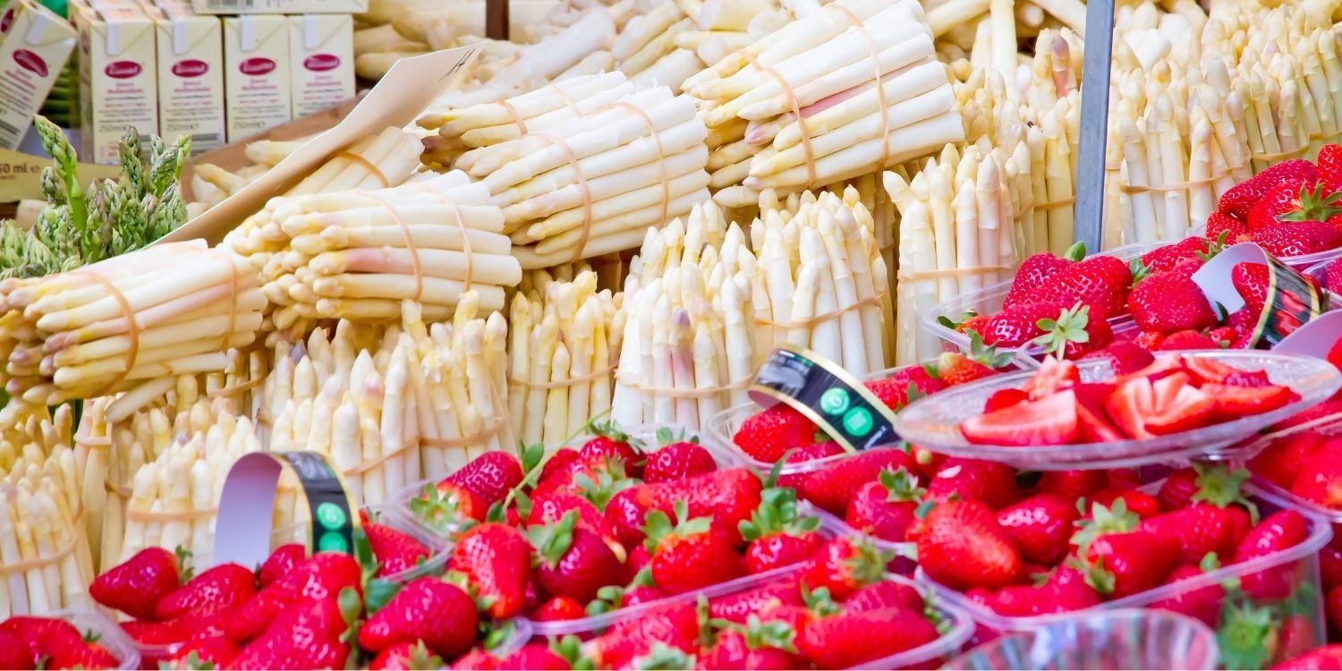 An einem Marktstand werden viele Bündel mit weißem Spargel und Körbchen mit Erdbeeren angeboten.. Artikel "Saisonal einkaufen liegt im Trend" öffnen.