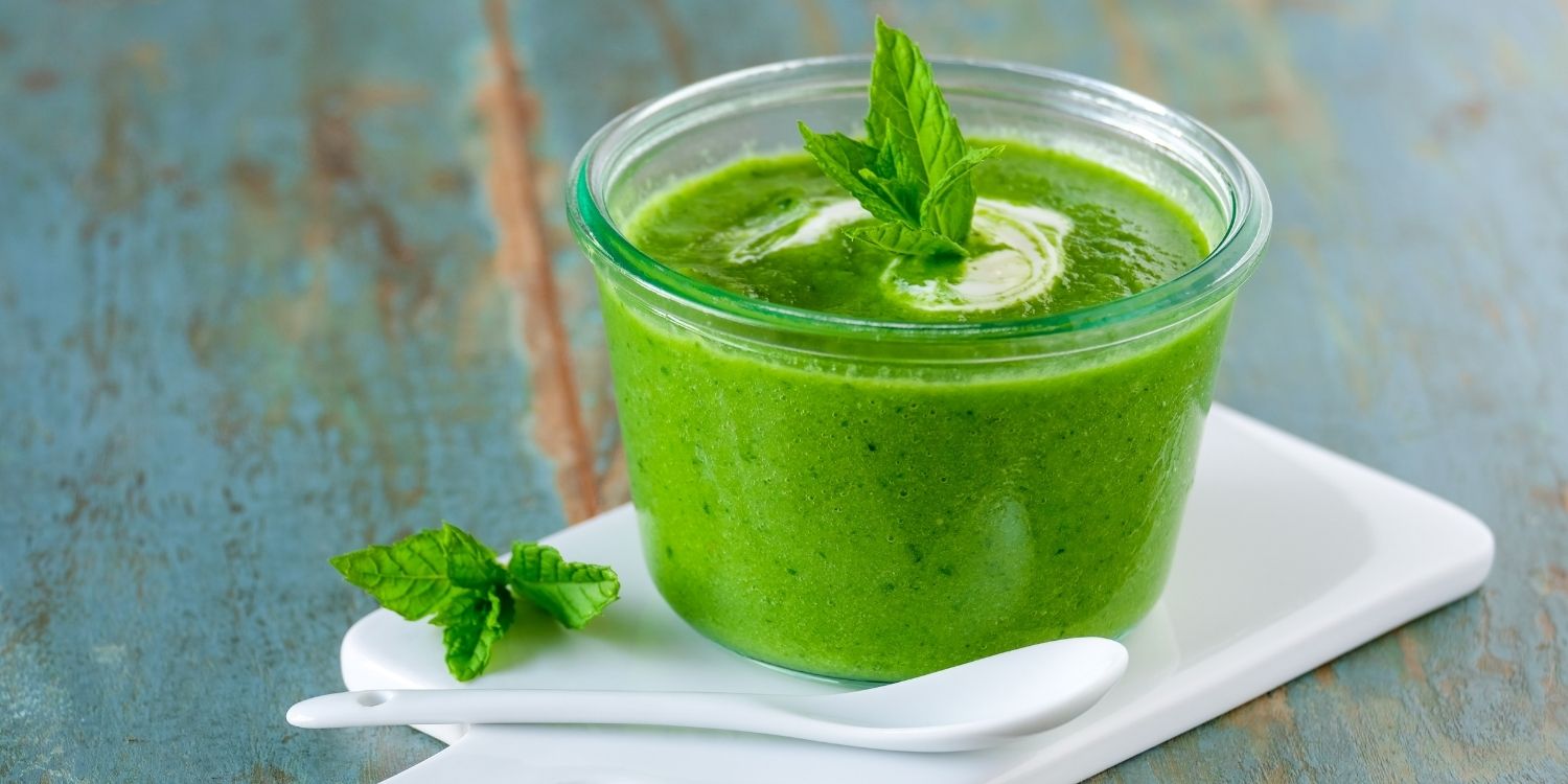 Auf einem weißen Brett steht eine Glasschüssel mit grüner Erbsensuppe, garniert mit Minzblättern. Daneben liegt ein Löffel.. Artikel "Gesundheitstrend: Scheinfasten" öffnen.