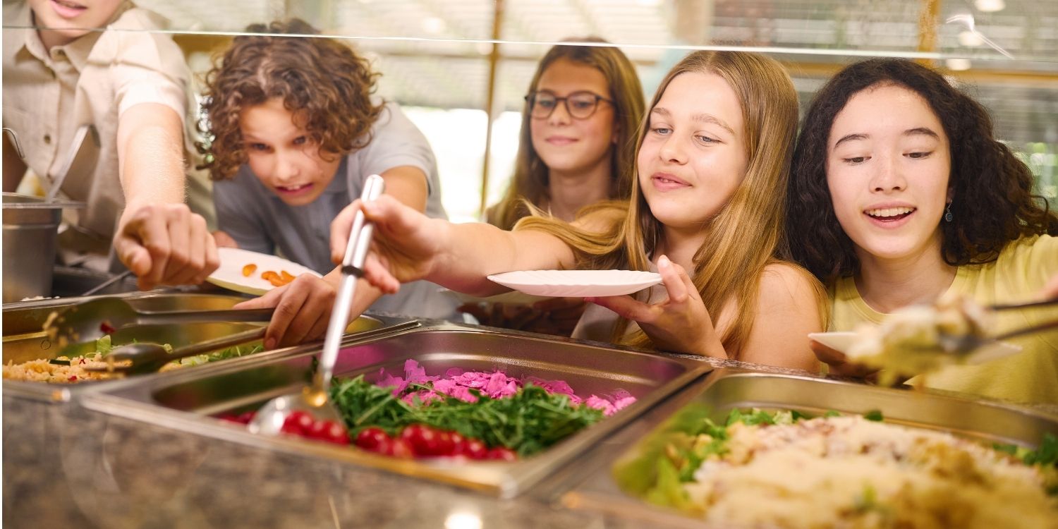 Eine Gruppe von Schülerinnen und Schülern lehnt sich über eine Essensausgabe und bedient sich mit Mittagessen.. Artikel "Neue WHO-Leitlinie zur Verbesserung des Schulessens" öffnen.