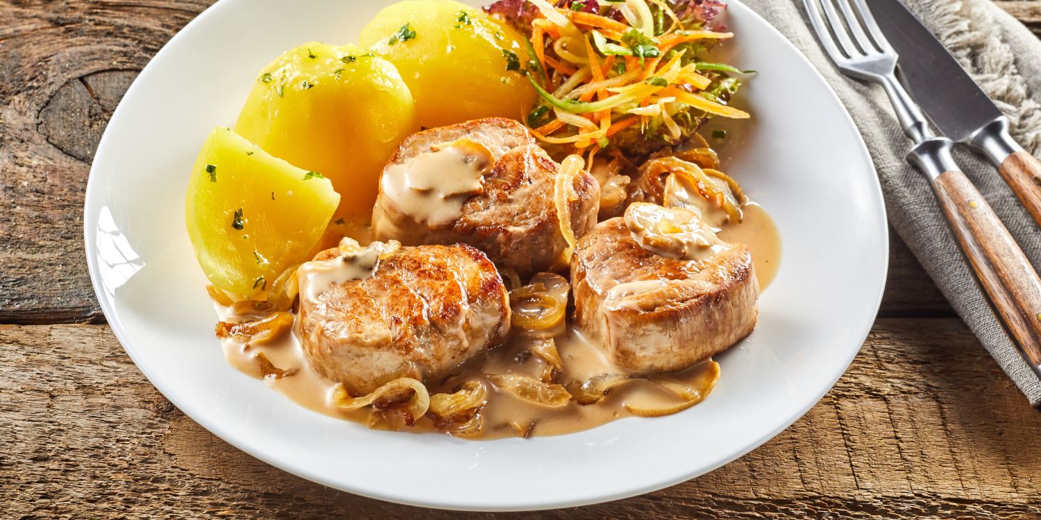Auf einem weißen Teller liegen drei Stücke Schweinefilet mit einer Zwiebelsoße und Kartoffeln.. Artikel "Aktuelle Zahlen zum Fleischverzehr in Deutschland " öffnen.