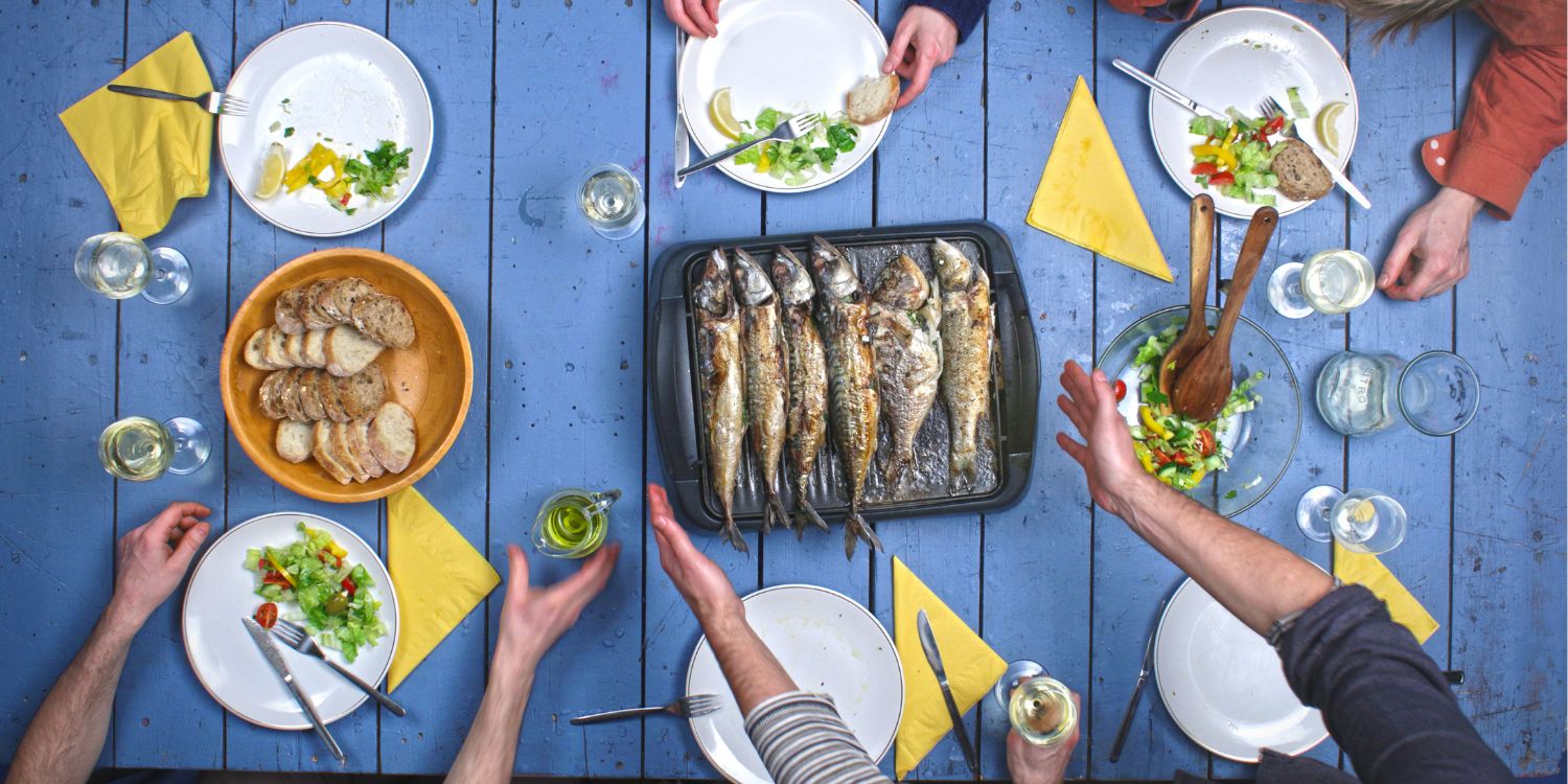 Auf einem blauen Holztisch liegt eine Platte mit gegrilltem Fisch, eine Salatschüssel und ein Korb mit Brot. Drumherum stehe weiße Teller und Gläser und Menschen greifen nach dem Essen.. Artikel "Gut mit Nährstoffen versorgt " öffnen.