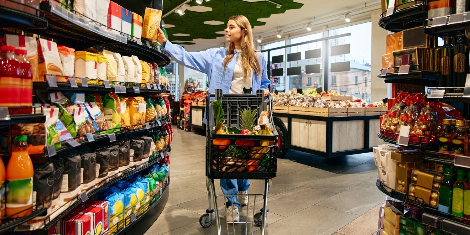 Eine junge blonde Frau in Jeans mit gefülltem Einkaufswagen nimmt eine Kuchenbackmischung aus einem Supermarktregal und betrachtet sie.. Artikel "Einkaufen für die Forschung" öffnen.