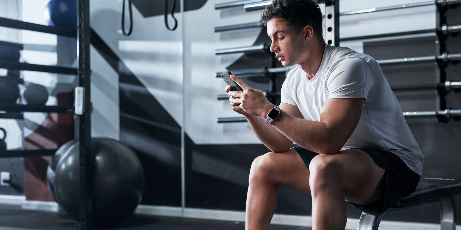 Ein junger Mann sitzt auf einem Trainingsgerät im Fitnessstudio und schaut besorgt auf sein Handy. . Artikel "Fitness-Content mit Nebenwirkungen" öffnen.