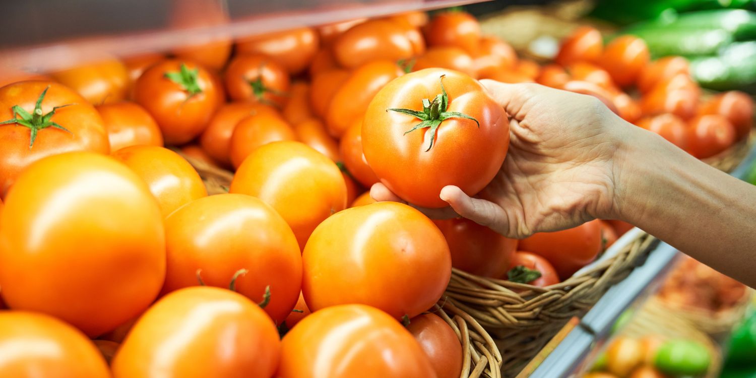Eine Hand greift eine Tomate aus einem Regal mit Tomaten in Gemüsekisten.. Artikel "Tomaten richtig lagern" öffnen.