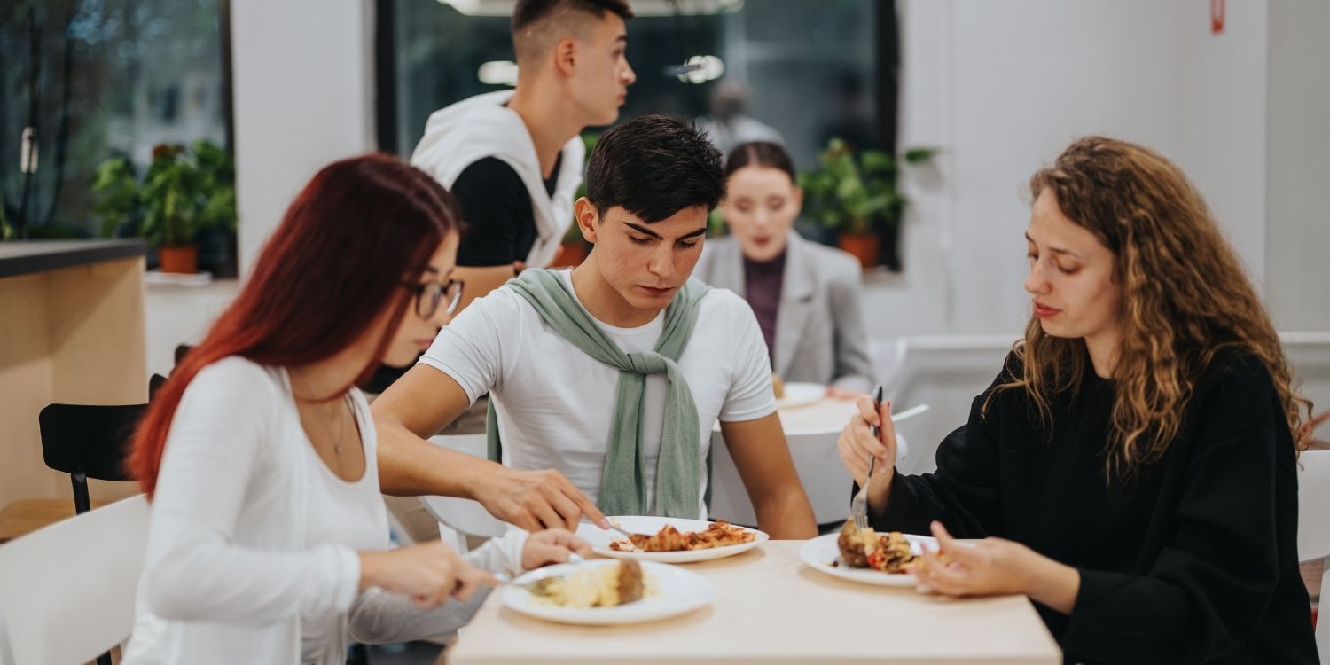  In einer Mensa sitzen zwei Studentinnen und ein Student gemeinsam beim Essen an einem Tisch.. Artikel "Deutlich mehr vegane Gerichte in deutschen Hochschulmensen" öffnen.