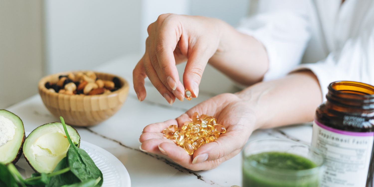 Auf einem weißen Küchentisch steht ein Körbchen mit Nüssen, ein grüner Smoothie, Salat und ein Glas mit Kapseln. Eine Frau hat viele Kapseln in ihrer Hand und nimmt eine davon.. Artikel "Vitamine – Viel hilft viel? " öffnen.