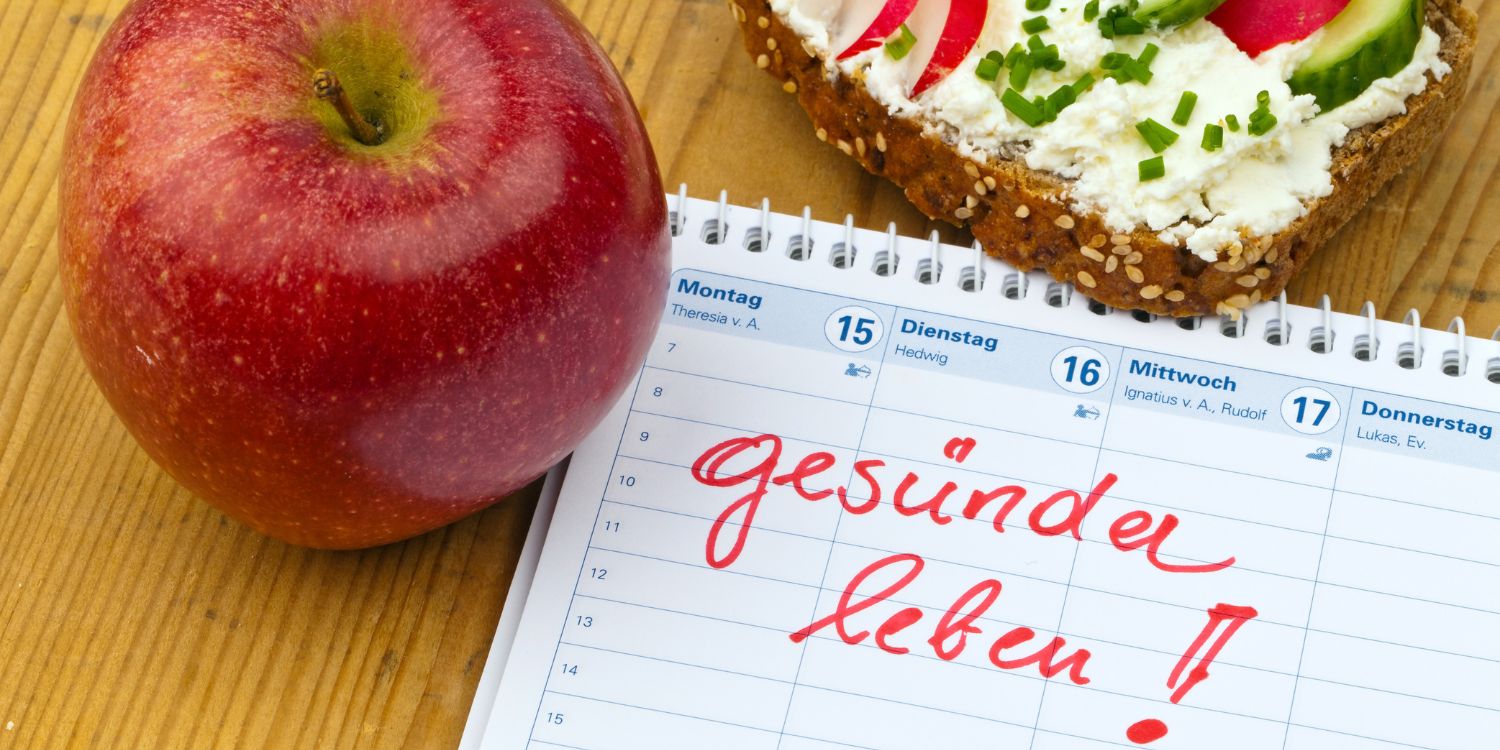 Auf einem Holztisch liegt ein Kalender mit dem Eintrag „Gesünder leben“, daneben ein roter Apfel und ein Vollkornbrot mit Frischkäse, Radieschen, Gurke und Schnittlauch belegt.. Artikel "Was hält uns davon ab, gesund zu leben?" öffnen.