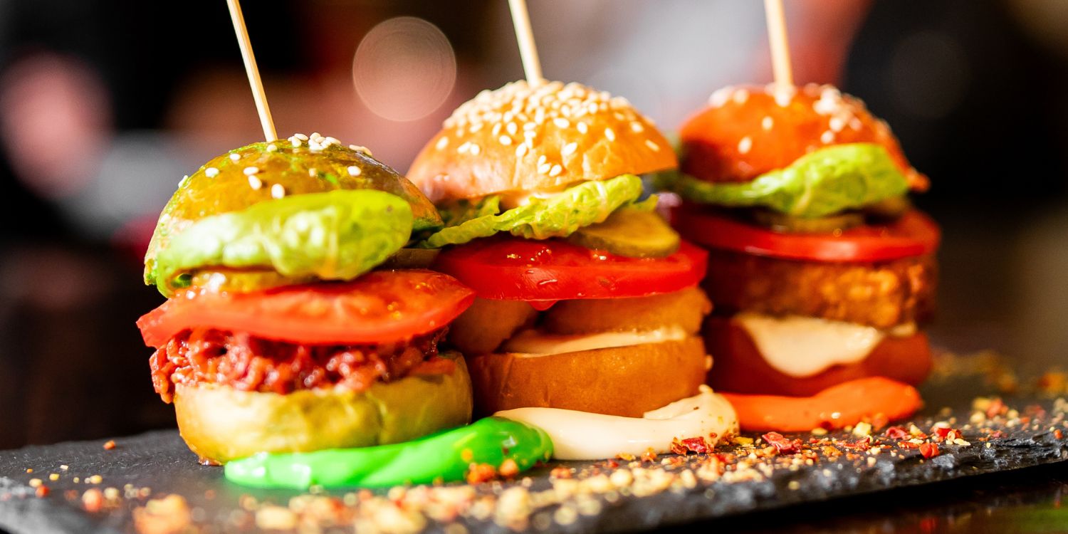 Auf einer dunklen Fläche stehen drei Miniburger mit verschiedenen Zutaten gefüllt und jeweils einem Holzspieß fixiert.. Artikel "USA auf dem Teller" öffnen.