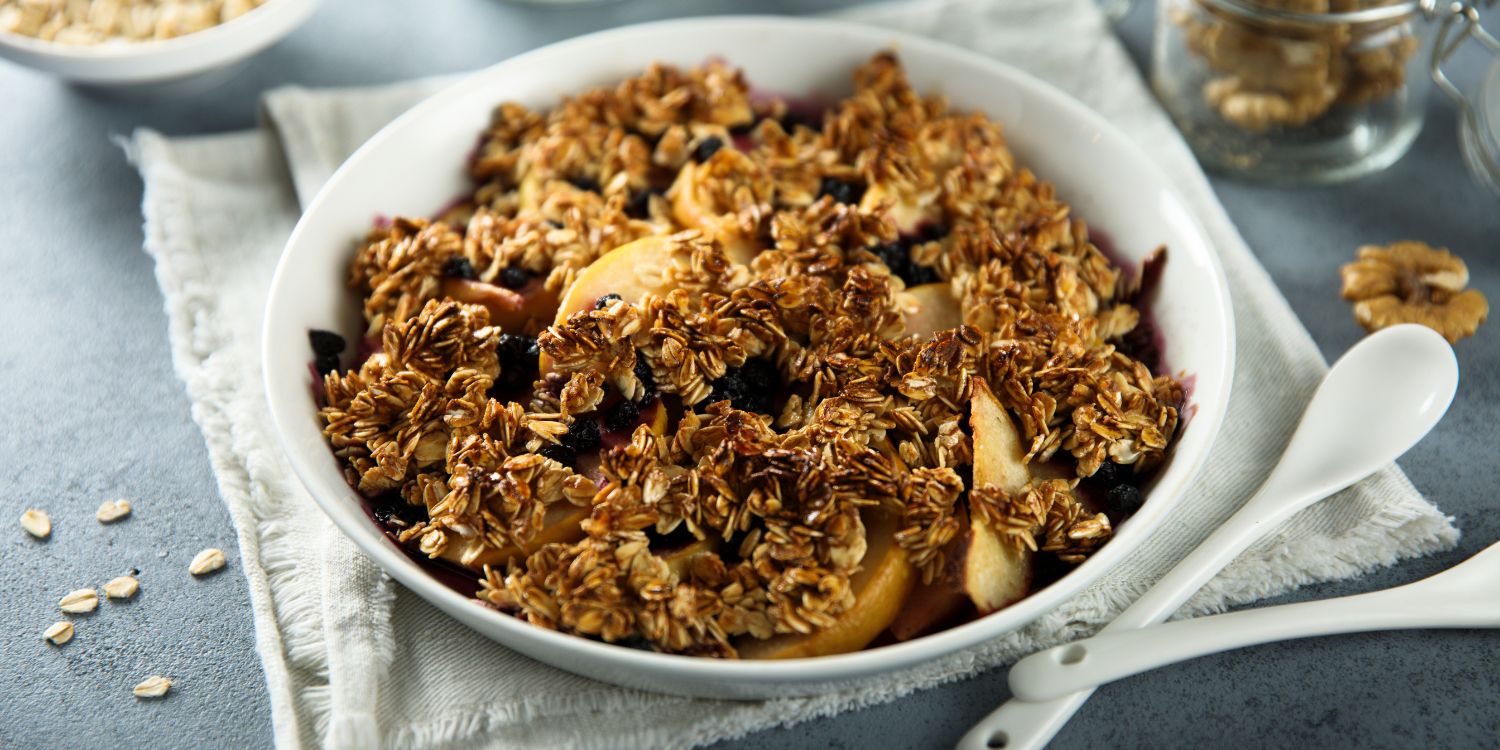 Auf einer dunklen Tischplatte steht eine weiße Schale mit gebackenen Haferflocken, Apfelspalten, Trockenfrüchten und Walnüssen. Daneben liegen ein Löffel und Walnusskerne.. Artikel "Warmes Frühstück für kalte Tage" öffnen.