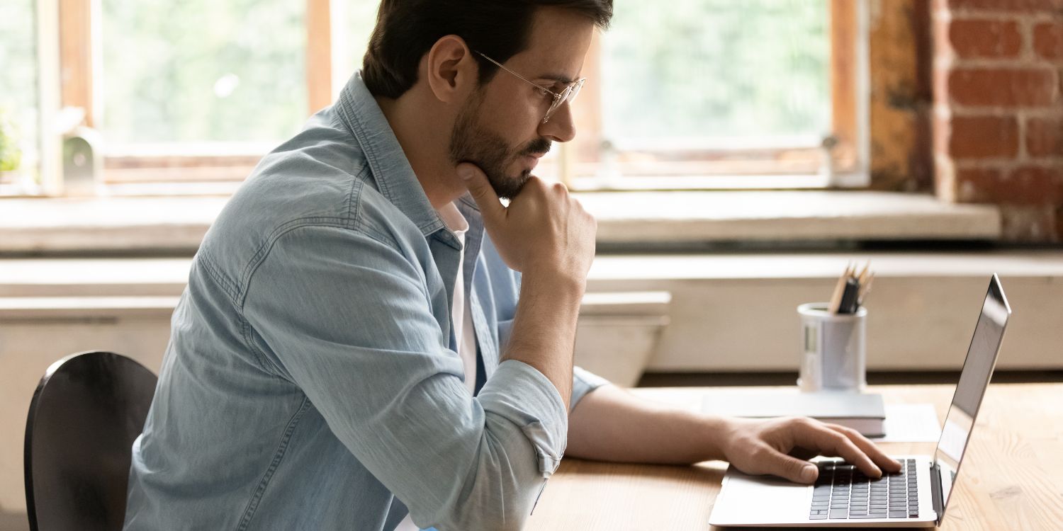 Ein dunkelhaariger Mann im hellblauen Hemd mit Bart und Brille sitzt konzentriert vor einem Laptop und stützt das Kinn in seine Hand.. Artikel "Kita- und Schulverpflegung professionell beschaffen" öffnen.
