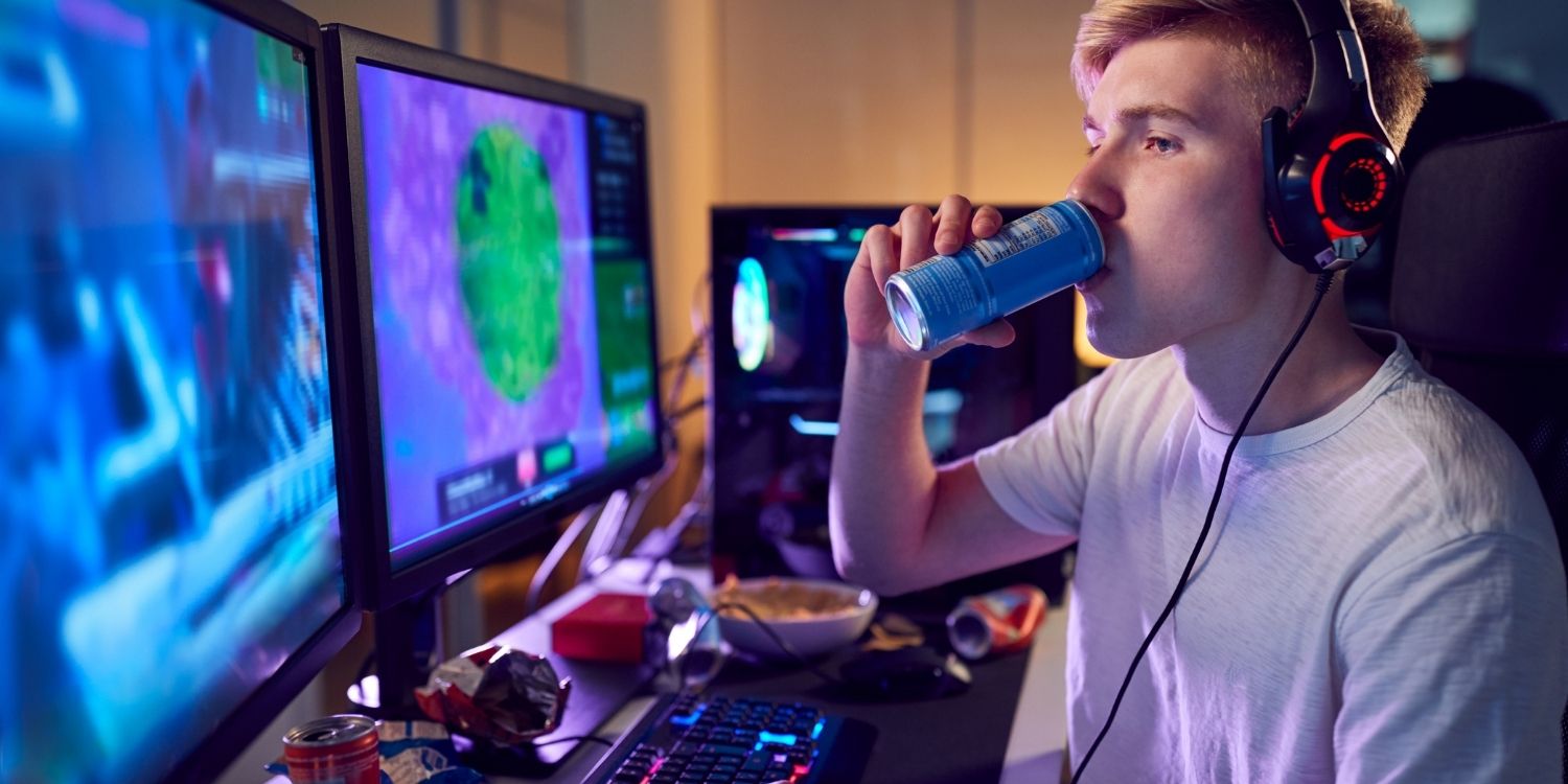 in blonder Jugendlicher im weißen T-Shirt mit Kopfhörern sitzt vor mehreren Bildschirmen und trinkt aus einer Dose.. Artikel "Lebensmittelwerbung auf Social Media erkennen" öffnen.