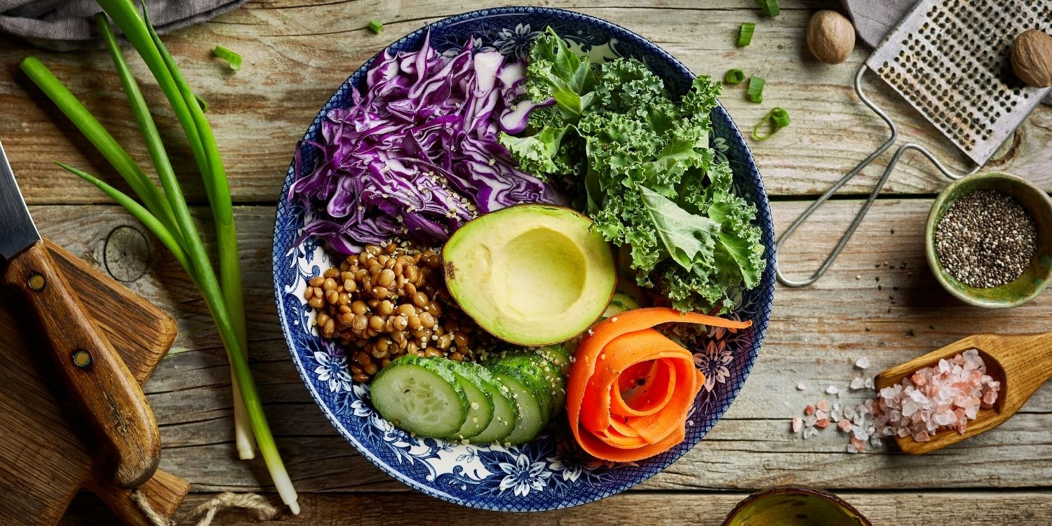 In einer blau-weiß gemusterten Schüssel sind Rotkohlstreifen, Blattsalat, Linsen, Salatgurke, Möhre und Avocado angerichtet. Auf dem Tisch daneben liegen Frühlingszwiebeln, Salz, Pfeffer und Muskatnuss.. Artikel "Bunte Winterbowls" öffnen.
