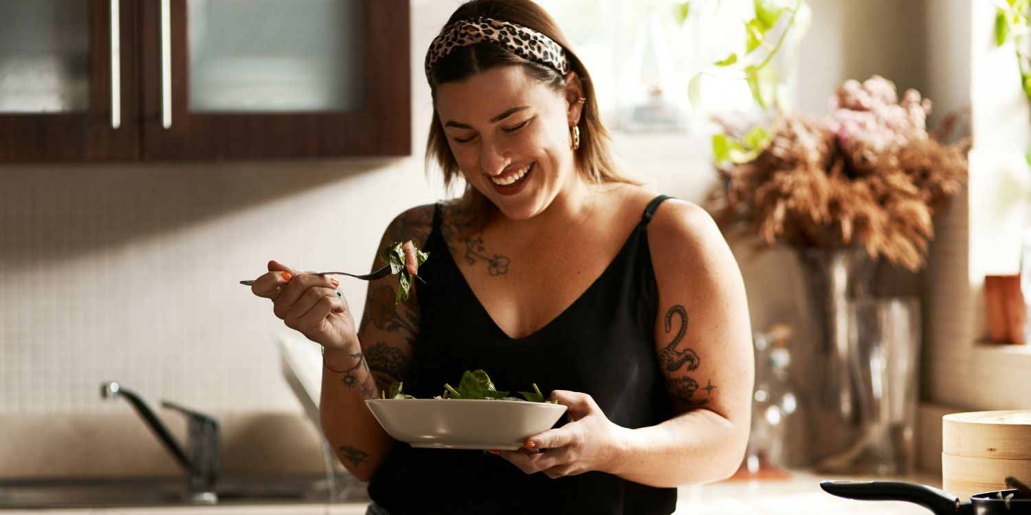 Eine junge dunkelhaarige Frau steht in einer Küche und isst Salat aus einem weißen Teller.. Artikel "Der Weg zum Wohlfühlgewicht" öffnen.