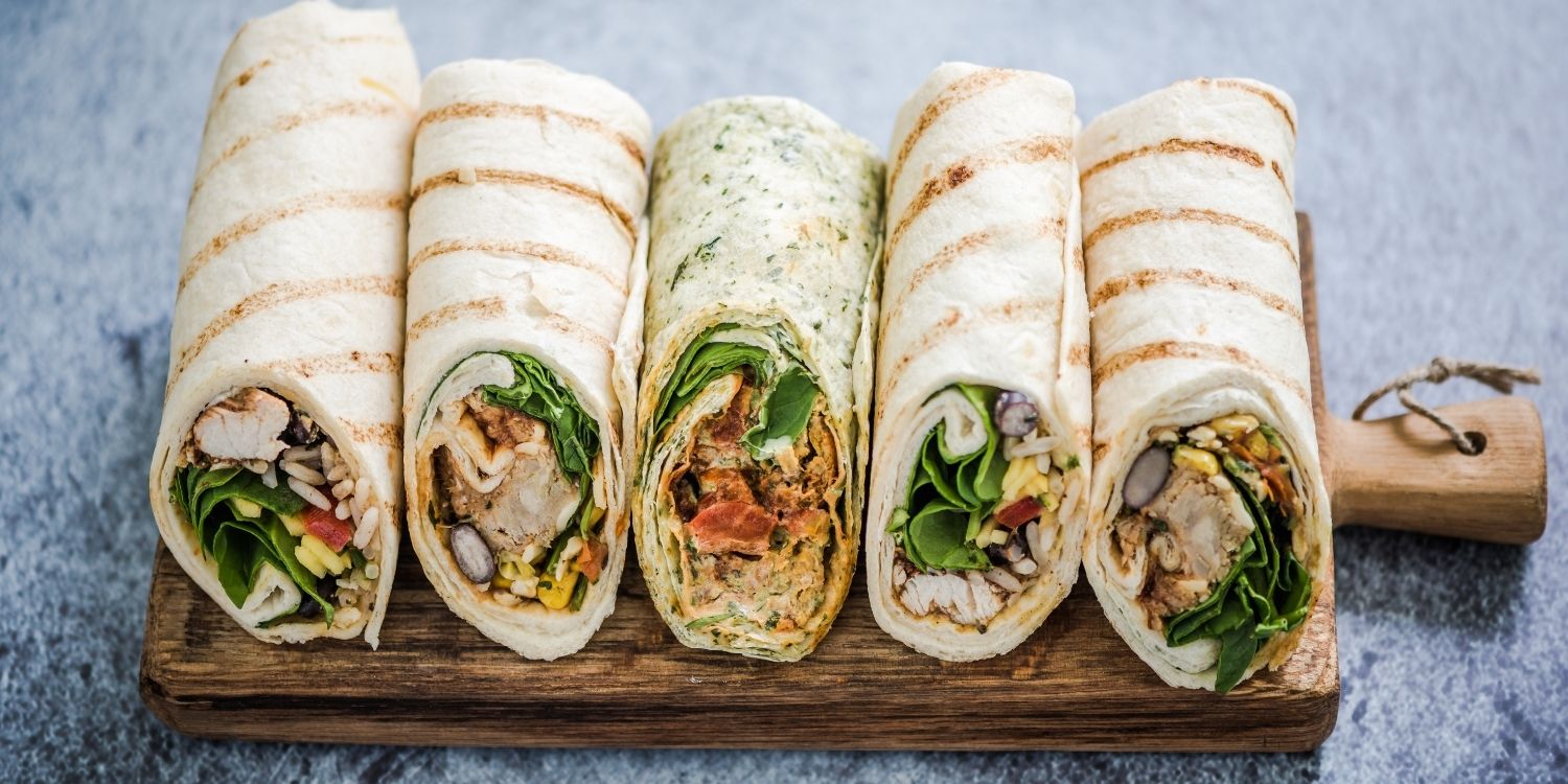 Auf einem Holzbrett liegen fünf Wraps nebeneinander, die unterschiedlich mit Salat, Gemüse, Fleisch und Reis gefüllt sind.. Artikel "Wraps selbst gefüllt" öffnen.