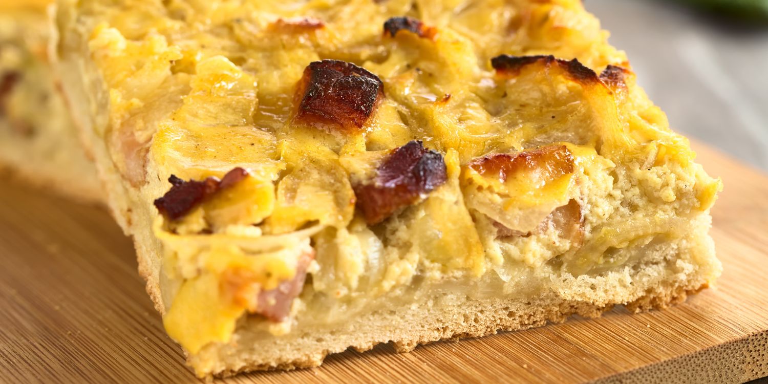 Auf einem Holzbrett liegt ein quadratisches Stück klassischer Zwiebelkuchen mit Speckstückchen.. Artikel "Lust auf Zwiebelkuchen" öffnen.