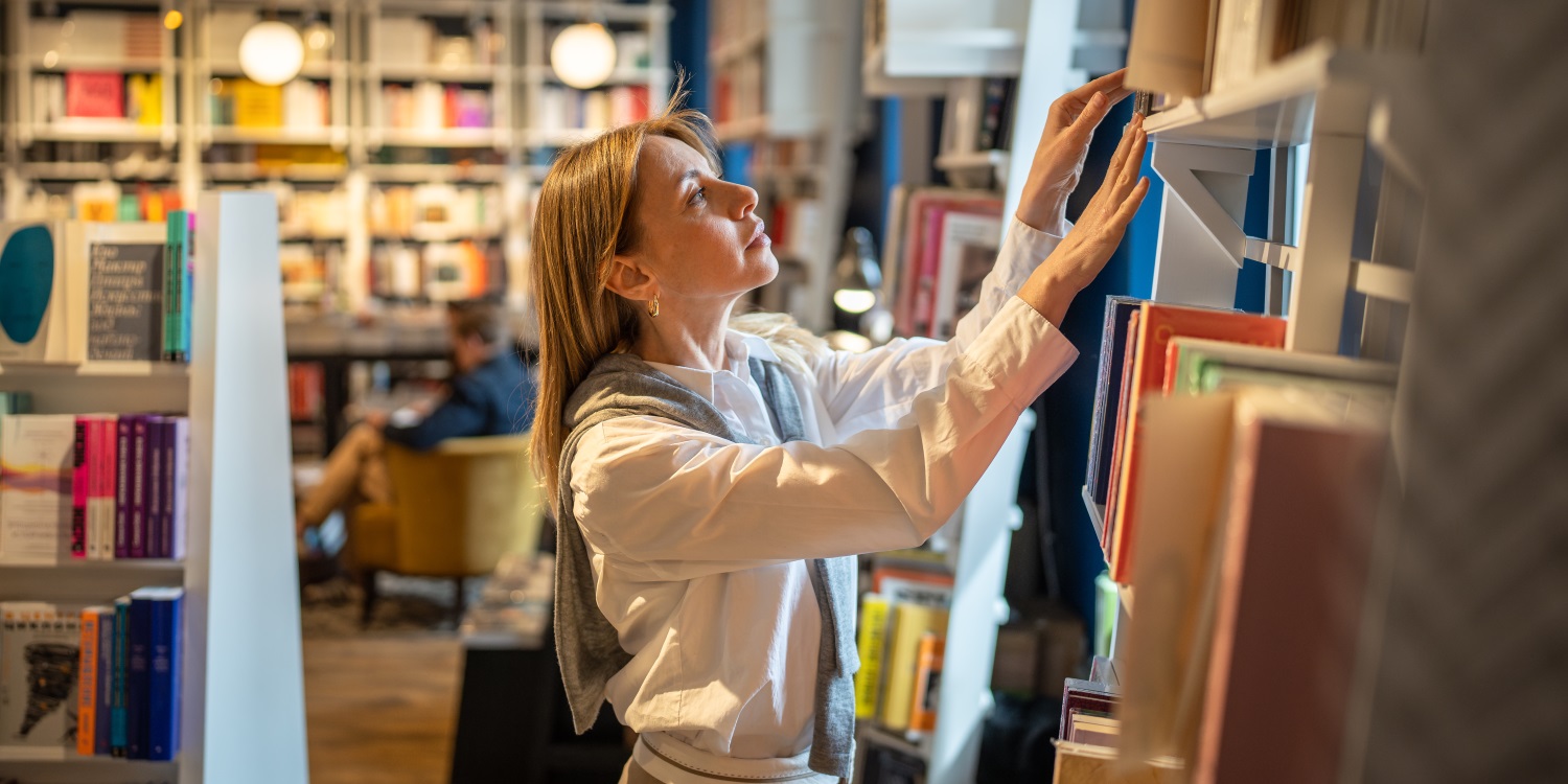Eine Frau steht vor einem Bücherregal und sucht sich ein Buch aus.. Artikel "Gute Unterrichtsmaterialien erkennen" öffnen.